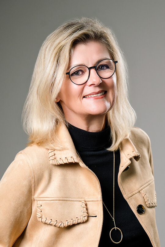 Lächelnde Frau mit blondem Haar, Brille, beiger Jacke.